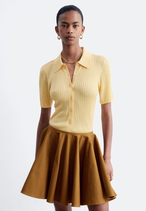 Femme portant une chemise jaune côtelée à manches courtes boutonnée et une jupe marron évasée, accessoirisée avec des boucles d'oreilles créoles dorées et un collier chaîne.