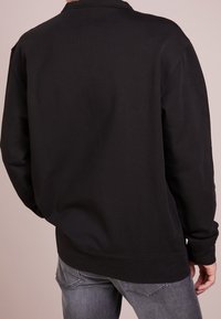 Sudadera de algodón negra con cuello redondo y mangas largas, que presenta un ajuste holgado y detalles de costura visibles. La vista posterior destaca un diseño simple.
