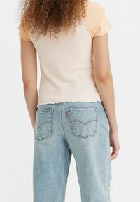 T-shirt med korte ærmer i ferskenfarvet foran og beige bagpå, med en scalloped kant, parret med lyseblå slidte denimjeans.