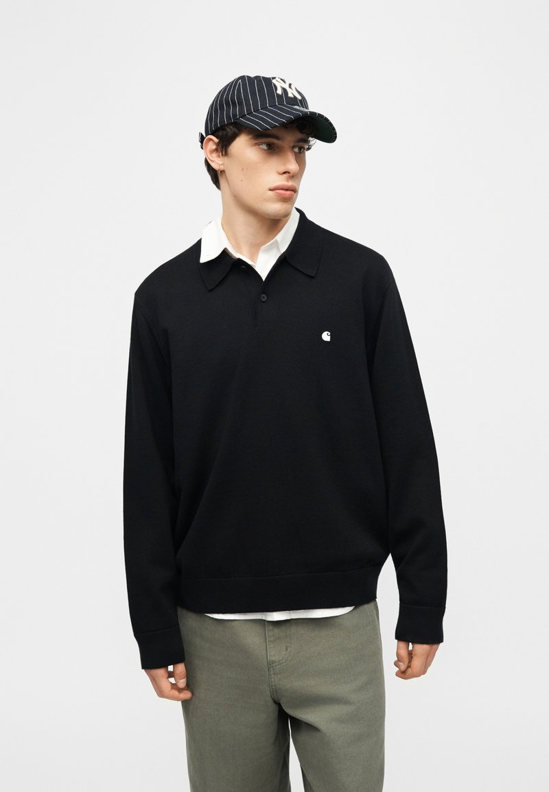 Junger Mann trägt ein schwarzes Polo mit langen Ärmeln, darunter ein weißes Hemd, olivfarbene Hose und eine marineblaue Mütze mit Nadelstreifen, blickt nach rechts.