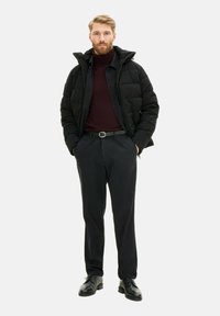 Zwarte gewatteerde jas over een bordeauxrode coltrui, gecombineerd met zwarte broek en schoenen. Voorzien van een ritssluiting en een riem. Vlakke textuur.