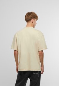 Beige oversized bomuld t-shirt med korte ærmer, rund hals og lige kant, båret bagfra. Stoffet virker blødt med minimal tekstur.