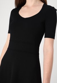 Černé pletené šaty s přiléhavým trupem, krátkými rukávy a kulatým výstřihem. Obsahují vodorovné švy v oblasti pasu. Měkká textura.