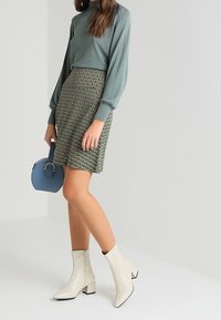 Un pull en tricot vert, une mini-jupe à motifs, des bottines blanches et un sac à main rond bleu. Textures lisses et formes structurées visibles.