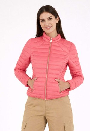 Donna sorridente che indossa una giacca trapuntata aderente rosa corallo con tasche con cerniera e pantaloni cargo beige, mani nelle tasche, in piedi su sfondo bianco.