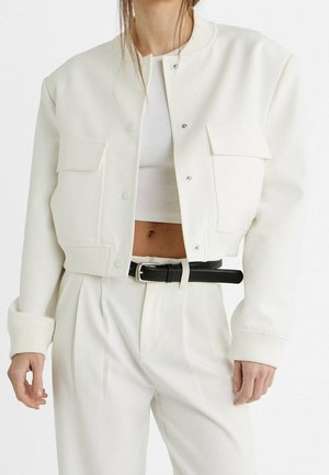 Femme portant une veste blanche courte à boutons-pression, un pantalon blanc taille haute, une ceinture noire et un crop top blanc côtelé.
