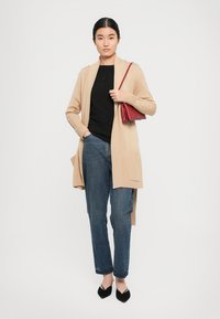 Beige neulecardigan avoimella edessä, raidoitetuilla hihoilla ja sivutaskuilla, yhdistetty mustan t-paidan ja sinisten suoranlahkeisten farkkujen kanssa. Punainen käsilaukku.