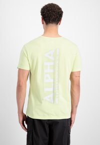 Svijetlozelena majica s kratkim rukavima s velikim bijelim natpisom "ALPHA INDUSTRIES" na leđima. Izrađena je od mekanog, teksturiranog pamučnog materijala.