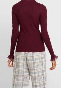 Bordeauxfarbener Rippstrickpullover mit langen Ärmeln und durchsichtigen, rüschengesäumten Bündchen, kombiniert mit karierten, weit geschnittenen Hosen in Weiß, Schwarz und Gelb.