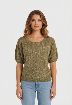 Donna che indossa un maglione a maniche corte lavorato a maglia di colore verde oliva e jeans azzurri chiari, in piedi contro uno sfondo chiaro e uniforme.