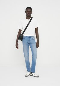 Ανοιχτό μπλε τζιν παντελόνι, λευκό μπλουζάκι, μαύρη τσάντα χιαστί και λευκά αθλητικά παπούτσια με μαύρες ρίγες. Νέος άνδρας που στέκεται μπροστά σε λευκό φόντο.