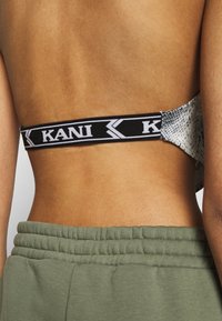 Černobílý elastický pásek s logem "KANI", spárovaný s látkou se vzorem hadí kůže. Viditelná pokožka a zelený elastický pas pod ním.