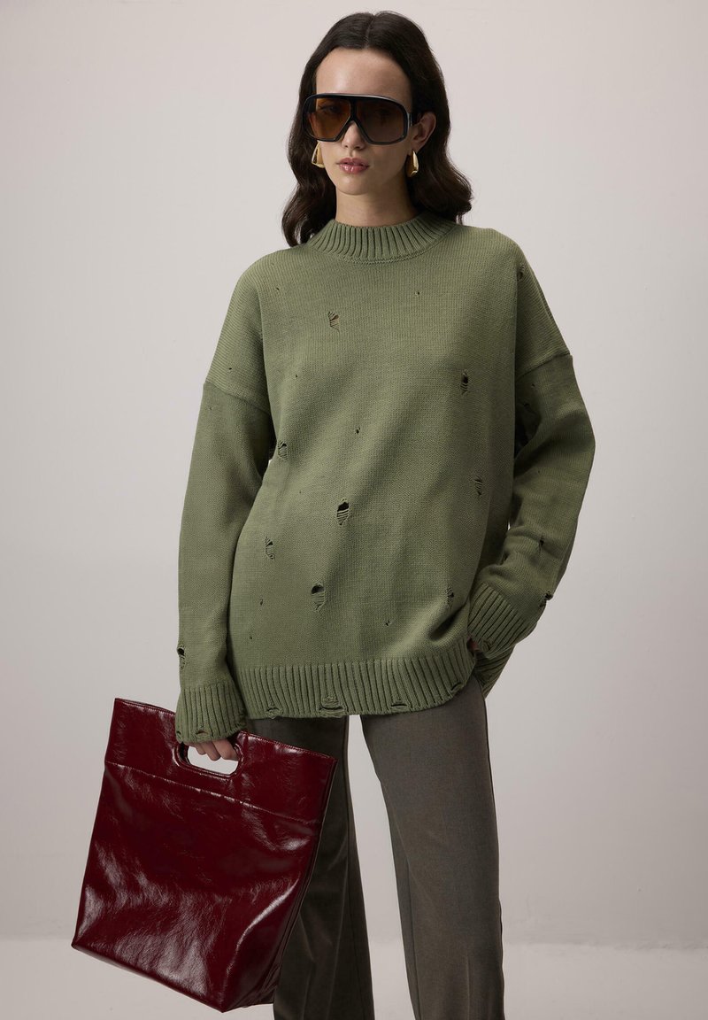 Oversized, oliwkowy sweter z postarzanymi detalami, prążkowanym kołnierzem i dołem. W zestawie z oliwkowymi spodniami i błyszczącą, burgundową torbą.