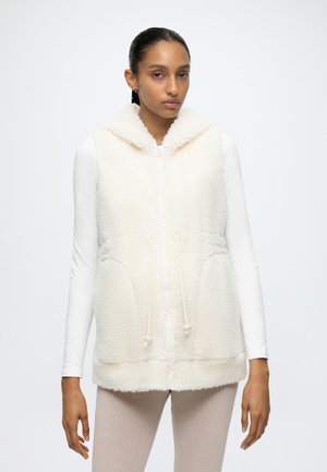 Femme portant une veste en polaire sans manches couleur crème avec une capuche, par-dessus une chemise blanche à manches longues et un pantalon beige clair, debout sur un fond blanc.