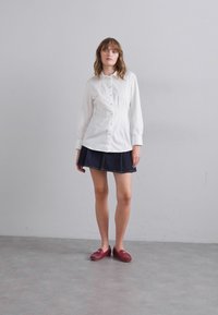 White button-up shirt with gathered details, paired with a dark denim skirt. Model wears red slip-on shoes, standing on a gray surface.