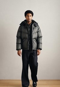 Puhasta tamnozelena jakna s kapuljačom, zatvaranjem na patentni zatvarač i velikim prošivenim dijelovima. Nosi se preko crne majice i uparen je s širokim crnim hlačama.