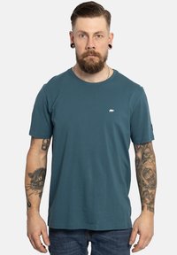 Teal crew neck t-shirt gemaakt van zachte stof, met een klein wit berenlogo op de borst en korte mouwen. Eenvoudig ontwerp, losse pasvorm.