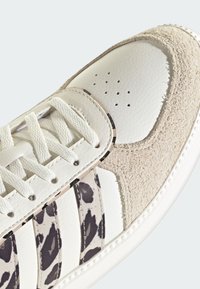 White sneaker with beige suede toe cap, perforated leather upper, white laces, and black-and-white spotted side stripes on a white sole.
