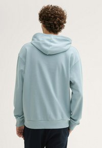 Helle blaue Kapuzenjacke mit weichem Material, ausgestattet mit einer großen Kapuze und gerippten Bündchen. Das Design umfasst einen lässigen Schnitt und einen sauberen, minimalistischen Stil.