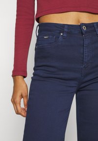 Tummansiniset farkut, joissa on slim fit -leikkaus, viisi taskua, huomaamaton ommel ja metallinappi kiinnitys. Yhdistetty tiukkaan punaisen pitkän hihan puseroon.