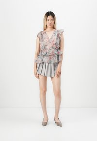 Blusa floreale trasparente con maniche a volant e peplum a strati; abbinata a una gonna grigia a vita alta con pieghe e tacchi neutri.
