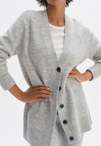 Šedý pletený kardigan s výstřihem do V, pěti černými knoflíky, žebrovanými manžetami a uvolněným střihem. Texturovaná tkanina s měkkým vzhledem.