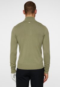 Grön stickad tröja med hög krage, ribbade manschetter och nederkant, med en liten logotyp på baksidan. Slät textur, figursydd design.