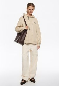 Sudadera beige con capucha y cordones, pantalones de pierna ancha color crema, y bolso tote marrón. La modelo lleva zapatos oscuros de deslizarse con puntas afiladas.