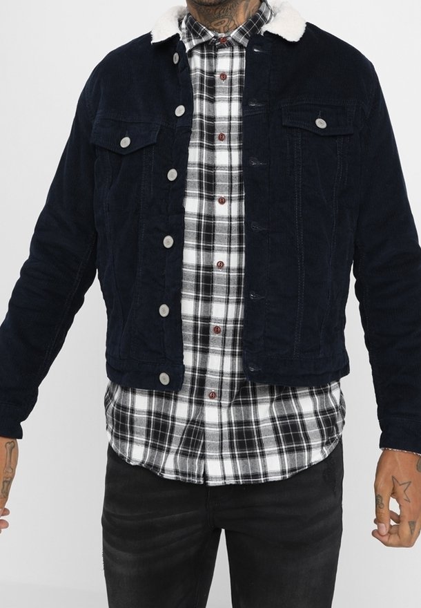 Mörkblå kordflorsjacka med vit fuskpälskrage, med knappstängning, två bröstfickor och en texturerad yta. Bärs över en rutigt skjorta.