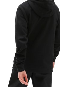 Persona che indossa una felpa nera con cappuccio e pantaloni neri, mostrata di spalle con una mano rilassata lungo il fianco.