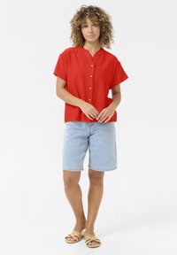 Rotes kurzärmliges Hemd mit Knopfleiste und gewelltem Kragen, kombiniert mit hellblauen Jeansshorts. Trägt beigefarbene Sandalen.