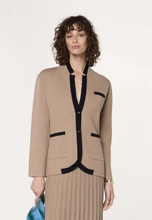 Vrouw met krullend haar draagt een beige vest met zwarte bies en knopen over een bijpassende geplooide rok, houdt een blauw patroonstof vast in haar linkerhand.