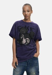 Fialová oversized tričko s veľkým čiernym grafickým motívom Mickey Mouse, s opotrebovanou textúrou a odvážnym písmom vpredu. Skombinované s modrými džínami.