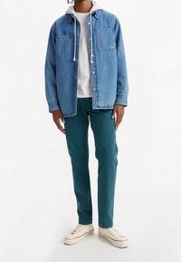 Camicia di jeans azzurro chiaro con due tasche sul petto, t-shirt bianca sotto, pantaloni turchesi e sneaker crema con dettagli rossi.