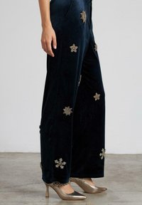 Pantalones de terciopelo navy de pierna ancha adornados con embellishments florales en oro y plata. Combinados con zapatos de tacón metálicos.