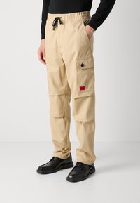 Beige cargo bukser med elastisk talje, snoretræk og sidelommer. Har en rød logo-patch og knapdetaljer på lommen.