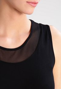 Top sin mangas negro con un escote de malla transparente, presentado con un cuello redondo; textura de tela suave.
