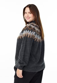 Nasmejana žena s dugom smešnom kosom koja nosi tamnosivi pulover s belim i narančastim geometrijskim uzorcima na ramenima, stoji naslonjena na belu pozadinu.