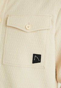 Poche de chemise en tissu beige texturé avec un rabat boutonné et une petite étiquette carrée noire présentant un motif abstrait blanc.