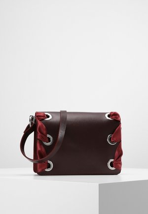 Dark brown leather handbag with silver grommets and red satin ribbon woven through corners, placed on a white pedestal.
