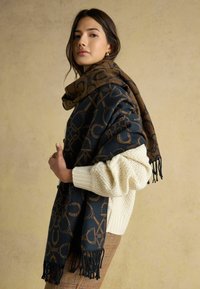 Veľká šál v tmavomodrej a hnedej farbe, s geometrickým vzorom, strapcami na okrajoch a mäkkou textúrou, nosená cez krémový sveter.