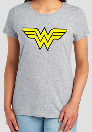 Woman wearing gray t-shirt with yellow Wonder Woman logo centered on the chest, paired with light blue jeans.