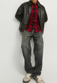 Chaqueta de cuero negra, camisa a cuadros roja y negra, jeans grises holgados, zapatillas beige con detalles negros, de pie contra un fondo neutro.