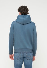 Μπλε φούτερ με κουκούλα, απαλό ύφασμα, άνετη εφαρμογή, ριμπ μανσέτες και τελείωμα, χωρίς εμφανή λογότυπα ή σχέδια, φορεμένο με ανοιχτό μπλε τζιν.