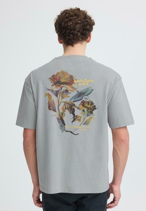Mann trägt ein graues T-Shirt mit buntem Blumen- und Insektenmuster sowie gelbem Schriftzug auf der Rückseite, steht vor einem schlichten Hintergrund.