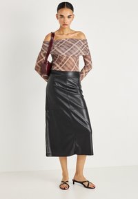 Jauna sieviete valkā apģērbu ar atklātiem pleciem un pledveida rakstu, melnu ādas vidēja garuma svārku, melnas papēžu sandales un pār plecu nēsā bordo krāsas somu.