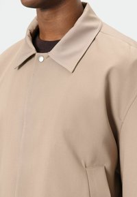 Lichtbeige jas met een spitse kraag, drukknoopsluiting en zijzak. Gladde textuur, minimalistisch ontwerp en getailleerde pasvorm.