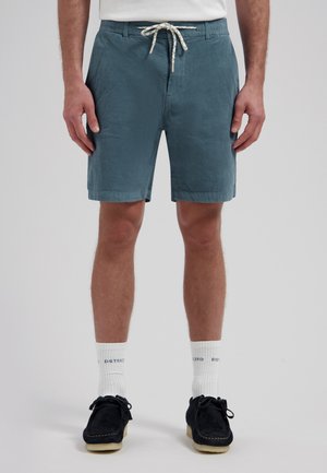 Man draagt teal kleurige ribfluwelen shorts met een witte trekkoord, witte merksokken en zwarte suède schoenen, staand tegen een grijze achtergrond.