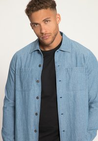 Lichtblauw denim shirt met een knoopsluiting aan de voorkant, twee borstzakken en een spread collar; gedragen over een zwart T-shirt.