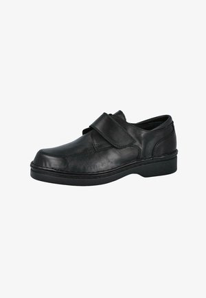 Zapato de cuero negro con puntera redondeada, cierre de velcro y suela baja de caucho. Presenta costuras a lo largo del borde y una textura suave.